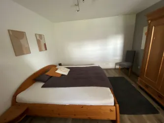 Ein gemütliches Schlafzimmer mit einem großen Bett und warmen Holzmöbeln. Die Wände sind hell, und es gibt einige dekorative Bilder sowie einen Stuhl in der Ecke.