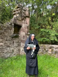 Eine Nonne steht vor einer alten Ruine im Wald. Sie hält ein Buch in der Hand und trägt eine lange schwarze Kutte.