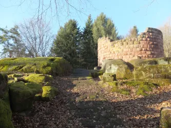 Eine alte Ruine aus Stein umgeben von moosbedeckten Felsen und Bäumen. Der Boden ist mit trockenen Blättern bedeckt und der Himmel ist klar.
