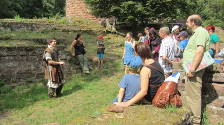 Eine Gruppe von Menschen hört einem Geschichtenerzähler in historischer Kleidung zu. Die Szene findet im Freien, umgeben von alten Ruinen, statt.