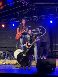 Zwei Musiker auf einer Bühne während eines Auftritts. Der Mann mit der Gitarre steht auf einem anderen Musiker, während sie Energien und Musik teilen.