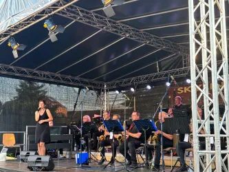 Eine Musikerin steht auf der Bühne und singt. Im Hintergrund spielt eine Band mit mehreren Musikern auf verschiedenen Instrumenten.