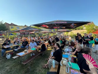 Eine fröhliche Gruppe von Menschen sitzt unter Sonnenschirmen auf Holzbänken. Es scheint eine Veranstaltung im Freien zu sein, mit vielen Gästen und einer entspannten Atmosphäre.