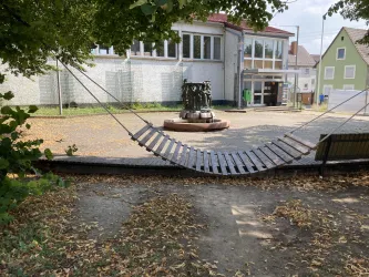 Eine Hängematte hängt zwischen Bäumen in einem ruhigen Platz. Im Hintergrund sind Gebäude und ein Brunnen zu sehen.