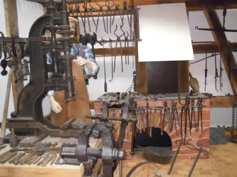 Eine Werkstatt mit traditionellen Schmiedewerkzeugen und einem großen Amboss. Im Hintergrund ist ein Schmied zu sehen, der an einem Projekt arbeitet.