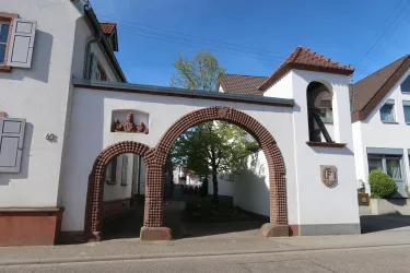 Ein historisches Tor mit einem runden Bogen und einer kleinen Uhr auf der Seite. Im Hintergrund sind Wohngebäude und ein Baum sichtbar.