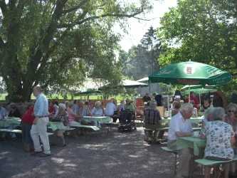 Eine lebhafte Gartenparty mit vielen Menschen, die an Tischen sitzen und sich unterhalten. Über den Tischen stehen Sonnenschirme und es ist eine freundliche Atmosphäre im Freien.
