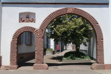 Ein historischer Durchgang mit zwei Bögen und dekorativen Elementen. Im Hintergrund ist ein grüner Baum und ein gepflasterter Weg sichtbar.