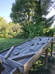 Ein großes, hölzernes Spielgerät steht im Freien, umgeben von Grün. Im Hintergrund ist ein Baum und ein gepflegter Garten sichtbar.