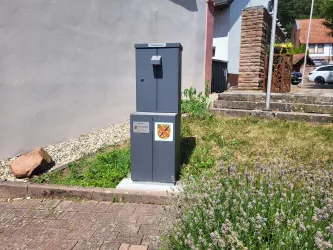 Ein grauer Trinkbrunnen steht auf einem gepflasterten Weg. Im Vordergrund sind grüne Pflanzen und Lavendel sichtbar.