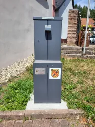 Ein grauer Trinkbrunnen steht auf einem Pflasterplatz in einer grünen Umgebung. Neben ihm befinden sich Steine und eine Wand im Hintergrund.