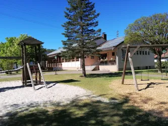Ein Spielplatz mit Klettergerüst, Schaukeln und Sandfläche. Im Hintergrund steht ein Gebäude und es ist schön sonnig.