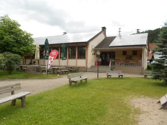 Ein gemütliches Gebäude mit mehreren Fenstern und Sonnenschirmen. Im Vordergrund sind Bänke auf einer Wiese und ein Eis-Stand.