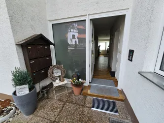 Ein einladender Eingangsbereich mit einer Haustür und einem kleinen Vorplatz. Neben der Tür steht ein Blumtopf und ein Briefkasten.