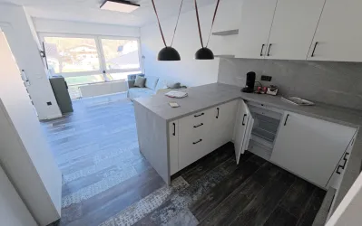 Eine modern eingerichtete Küche mit hellen Schränken und einem großzügigen Tisch. Im Hintergrund ist ein gemütlicher Wohnbereich mit Fenster und Tageslicht zu sehen.