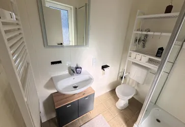 Ein modernes Badezimmer mit einem Waschbecken, einem Spiegel und einer Dusche. Es enthält auch Regale für Badutensilien und Handtücher.