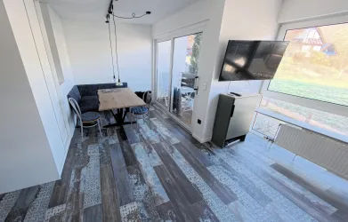 Ein modernes Wohnzimmer mit einem Esstisch und Sesseln. Große Fenster bieten viel Licht und einen Blick nach draußen.