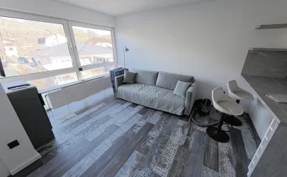 Ein modernes Wohnzimmer mit einem grauen Sofa und zwei weißen Barhockern. Große Fenster bieten einen Blick auf die Umgebung und sorgen für viel Tageslicht.