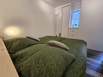 Ein gemütliches Schlafzimmer mit zwei großen Betten, die mit grünen Bettdecken bedeckt sind. Ein Fenster lässt sanftes Licht herein und eine Kommode ist im Hintergrund sichtbar.