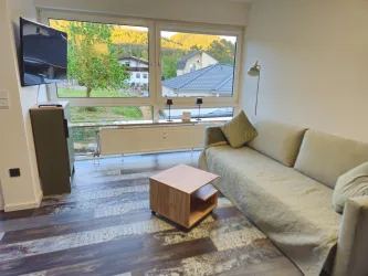 Ein gemütliches Wohnzimmer mit einem bequemen Sofa und einem kleinen Holzkaffee-Tisch. Große Fenster bieten einen Blick auf die umliegende Natur und die Nachbarhäuser.