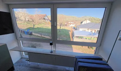 Ein Blick aus dem Fenster auf eine ruhige Wohnstraße mit mehreren Häusern und grünen Wiesen. Die Sonne scheint und der Himmel ist klar.