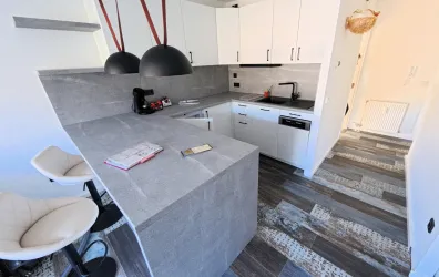 Eine moderne Küche mit einer grauen Arbeitsfläche und weißen Schränken. Im Hintergrund sind elegante Beleuchtung und Holzfußboden sichtbar.