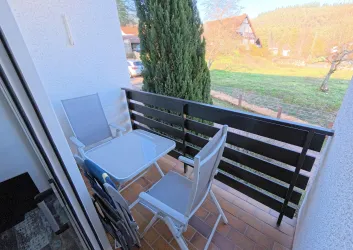 Ein kleiner Balkon mit einem Tisch und zwei Stühlen. Im Hintergrund ist eine grüne Wiese und einige Bäume zu sehen.