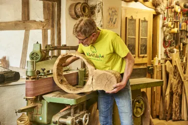 Ein Mann in einem gelben T-Shirt arbeitet in einer Holzwerkstatt. Er hält ein kunstvoll geformtes Holzstück und steht vor einer Holzmaschine.
