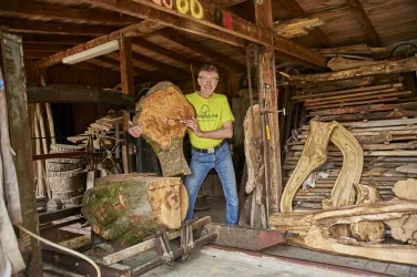 Ein Mann steht in einer Werkstatt mit einem großen Holzstück. Im Hintergrund sind weitere Holzstücke und Werkzeuge sichtbar.