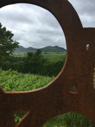 Ein runder Ausblick durch eine metallene Skulptur auf eine grüne Landschaft mit Hügeln im Hintergrund. Der Himmel ist bewölkt und verleiht der Szene eine ruhige Stimmung.
