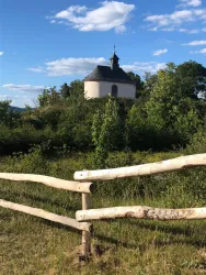 Eine kleine Kapelle steht auf einem Hügel, umgeben von Bäumen und Sträuchern. Im Vordergrund ist ein Holzzaun zu sehen, der die Landschaft umgibt.