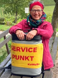 Eine lächelnde Frau sitzt auf einer Bank und hält eine Tasche mit der Aufschrift „SERVICE PUNKT“. Im Hintergrund sind grüne Weinberge sichtbar.