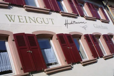 Ein Weingut mit dem Namen "Hechtmann" ist an einer Wand zu sehen. Die Fenster haben rote Fensterläden und weiße Vorhänge.