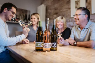 Eine gesellige Runde von vier Personen genießt Wein an einem Holzschirm. Im Vordergrund stehen drei Flaschen Wein, während die Gäste fröhlich anstoßen.