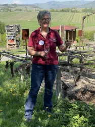 Eine Frau steht in einem Weinberg und hält ein Weinglas in der Hand. Im Hintergrund sind grüne Weinreben und sanfte Hügel zu sehen.