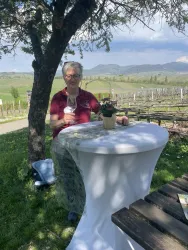 Eine Person sitzt an einem Tisch im Freien, umgeben von Weinbergen. Im Hintergrund sind sanfte Hügel und ein klarer Himmel zu sehen.