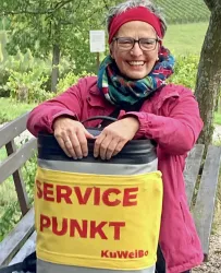 Eine lächelnde Frau sitzt auf einer Bank mit einem Servicepunkt neben sich. Sie trägt eine bunte Halstuch und eine Jacke.