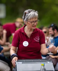 Eine ältere Dame in einem roten Polohemd serviert Getränke bei einer Veranstaltung im Freien. Im Hintergrund sind viele Menschen, die miteinander reden und die Atmosphäre genießen.