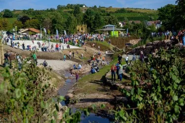 Eine große Menschenmenge versammelt sich in einer malerischen Landschaft mit Hügeln und einem kleinen Gewässer. Die Szene vermittelt eine lebhafte Atmosphäre und Naturverbundenheit.