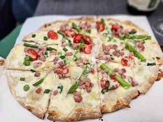 Eine leckere Pizza mit frischen Tomaten, grünem Spargel und Schinkenstückchen. Die Pizza ist in acht Stücke geschnitten und auf einem weißen Pizzateller serviert.