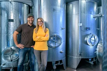 Zwei Personen stehen lächelnd vor großen, silbernen Weintanks. Die Umgebung wirkt modern und industriell.