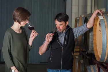 Ein Mann und eine Frau verkosten Rotwein in einem Weinkeller. Im Hintergrund sind Holzfässer zu sehen.