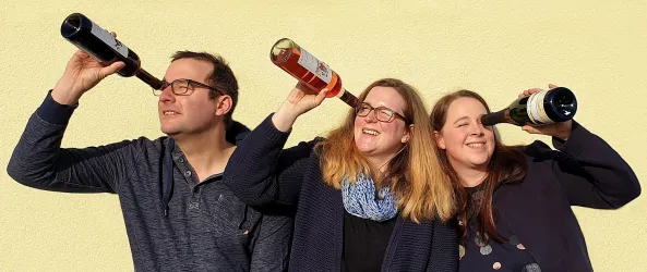 Drei Personen halten Weinflaschen ans Ohr, als ob sie lauschen. Sie stehen vor einer gelben Wand und lächeln.