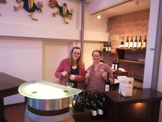 Zwei Frauen stehen an einer Weinbar und halten Flaschen Wein in den Händen. Im Hintergrund sind mehrere Weinflaschen auf Regalen zu sehen.