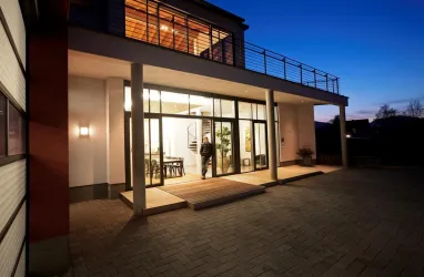 Ein modernes Haus mit großen Fenstern und einem Holzsteg. Die Szene ist bei Dämmerung aufgenommen, mit warmem Licht im Inneren.
