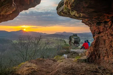 Eine beeindruckende Landschaft mit Sonnenuntergang hinter den Bergen. Zwei Personen sitzen im Vordergrund und genießen die Aussicht aus einer Höhle.