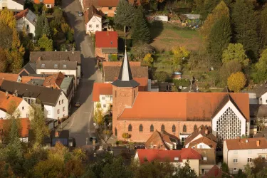 Eine Ansicht eines kleinen Dorfes mit Häusern und einer Kirche. Die Kirche hat einen markanten Turm und ist von bunten Bäumen umgeben.