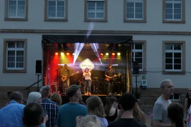 Auf dem Dorfplatz von Ruppertsberg stehen Menschen vor einer Bühne auf der eine Band Livemusik spielt. 