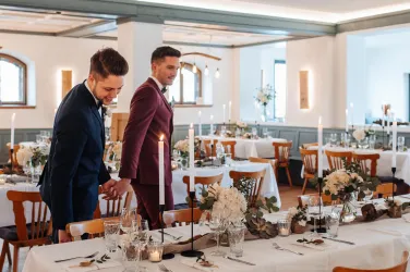 Zwei elegante Männer stehen an einem festlich gedeckten Tisch in einem schön dekorierten Raum. Kerzen und Blumen verleihen der Atmosphäre eine romantische Stimmung.
