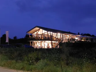 Ein modernes Gebäude am Abend, das von warmem Licht erleuchtet wird. Umgeben von Wiese und mit einem dunklen Himmel im Hintergrund.